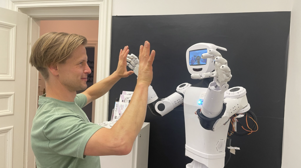 Mann mit grünem T-Shirt hebt die Hände vor einen weißen humanoiden Roboter mit Bildschirm als Kopf