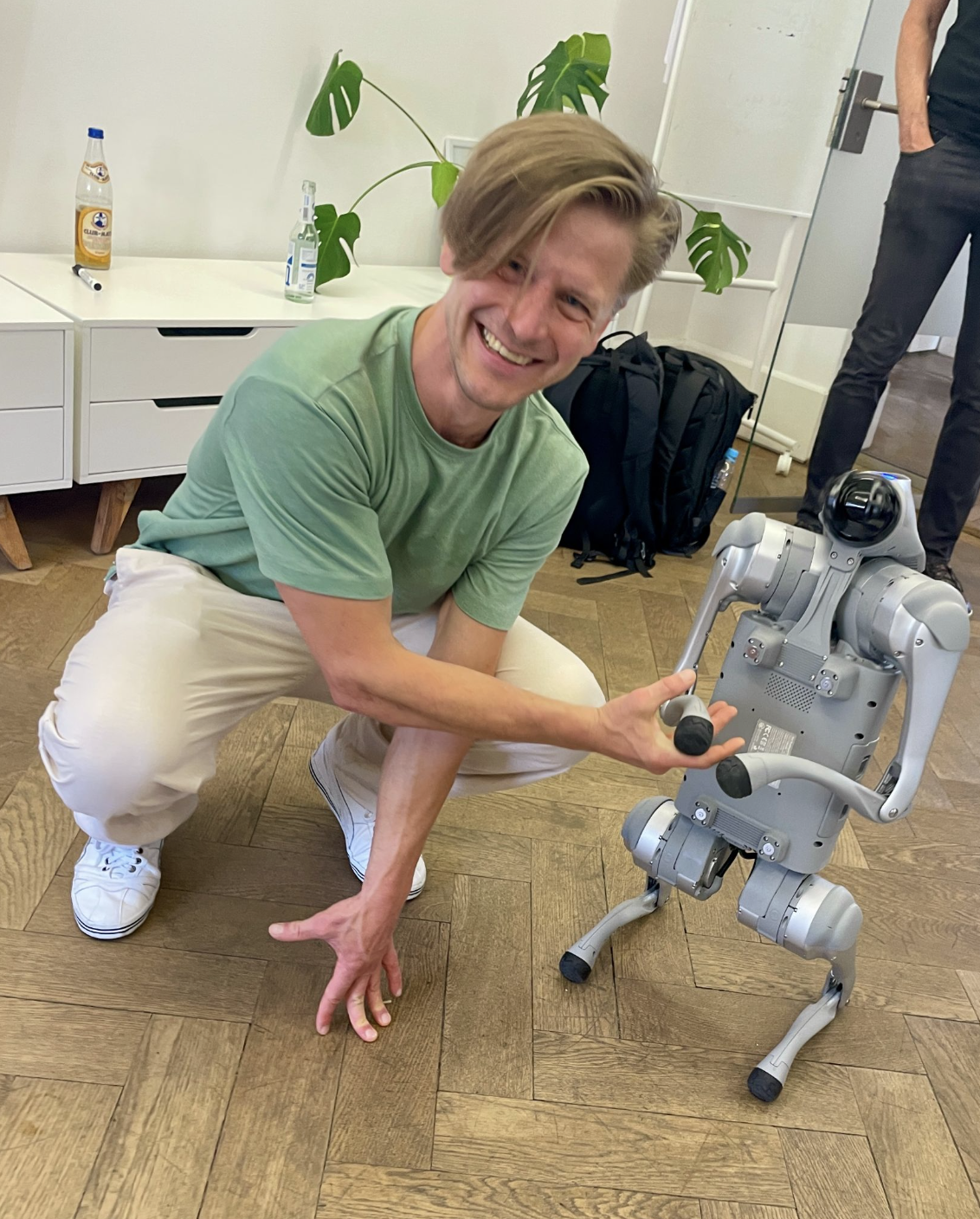 Junger Mann in grünem T-Shirt und weißer Hose hockt lachend neben einem kleinen humanoiden Roboter auf dem Holzboden
